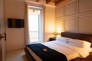 A hotel room with a bed, a TV mounted on the wall, a nightstand with a lamp, and a window with curtains. - Magnificent Residence overlooking Piazza Erbe (Verona)
