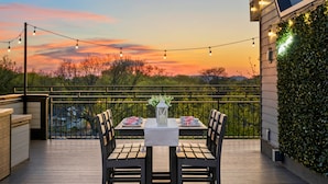 Um deck externo com uma mesa posta para dois, luzes de corda e vista para o pôr do sol.