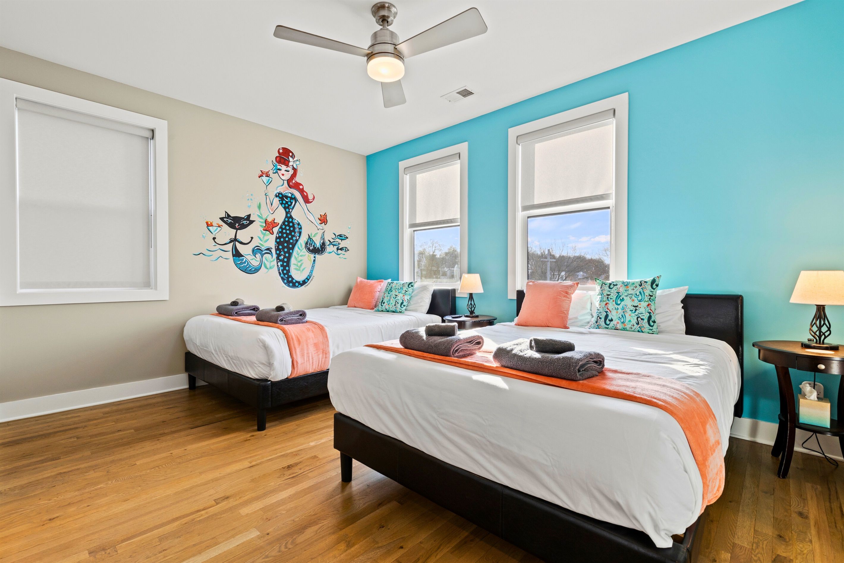 A hotel room with two beds, a ceiling fan, a mural, and large windows.