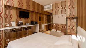 Habitación de hotel con cama, televisor, escritorio con cafetera y zona de estar con sillón y una mesita.