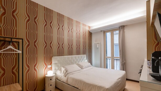 Habitación de hotel con cama, mesita de noche con lámpara, perchero y ventana con cortinas.