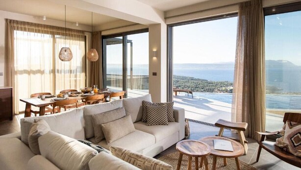 Un soggiorno ampio con un divano grande, un tavolo da pranzo e sedie. Grandi finestre offrono vista sul mare e sulla città.