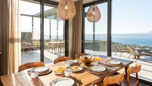 Zona pranzo con tavolo apparecchiato per la colazione, ampie finestre vista mare e lampade in vimini appese.