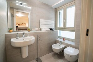 Une salle de bain moderne dotée d’un grand lavabo, d’un bidet et de toilettes, d’un miroir et d’une fenêtre laissant entrer la lumière naturelle.
