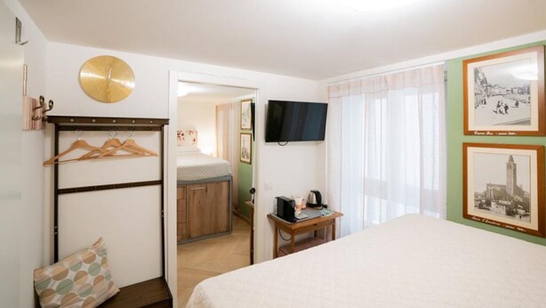 Habitación de hotel con cama, escritorio con computadora, televisor, perchero y reloj.