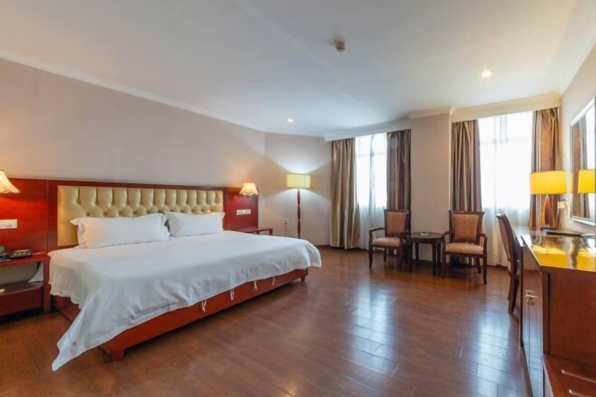 A hotel room with a large bed, a desk, two chairs, a lamp, and a window with curtains.