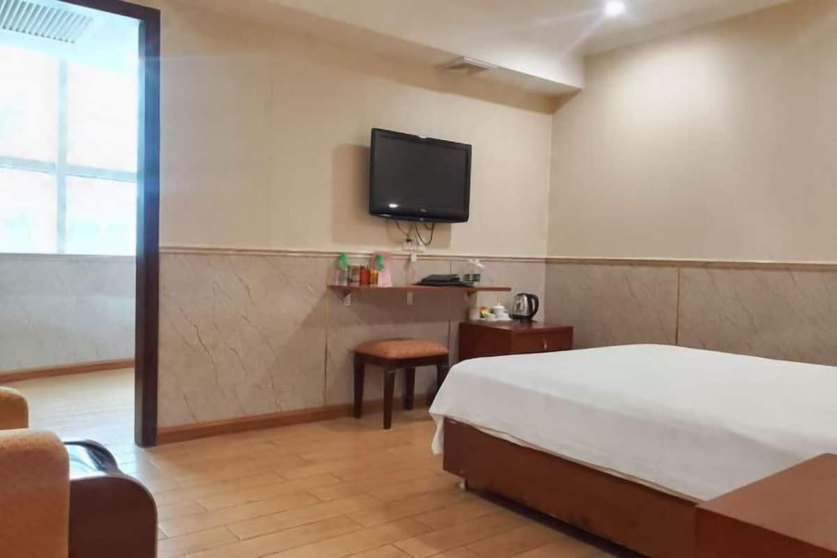 A hotel room with a bed, a television mounted on the wall, a small table with a kettle, and a window providing natural light.
