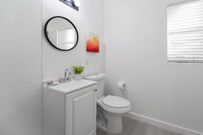 Uma casa de banho moderna com uma sanita, uma pia branca, um espelho e uma janela com persianas.