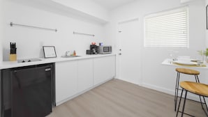 A modern kitchen with white cabinets, a dishwasher, a microwave, and a coffee maker. There is a window with blinds and a small table with stools. - Miami in Style Near Wynwood - We Host (Miami)