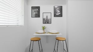 A dining area with a white table set for two, framed pictures on the wall, and a window with blinds. - Miami in Style Near Wynwood - We Host (Miami)