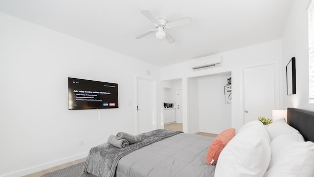 A modern bedroom with a bed, a ceiling fan, a TV mounted on the wall, and a kitchen area visible through an open door. - Miami in Style Near Wynwood - We Host (Miami)