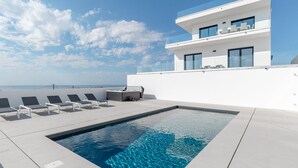 A modern rooftop pool area with a clear view of the ocean, a two-story building with large windows, and a row of sun loungers.