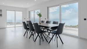 A modern dining area with a large table and chairs, large windows with a view of the sea, and a decorative vase on the table.