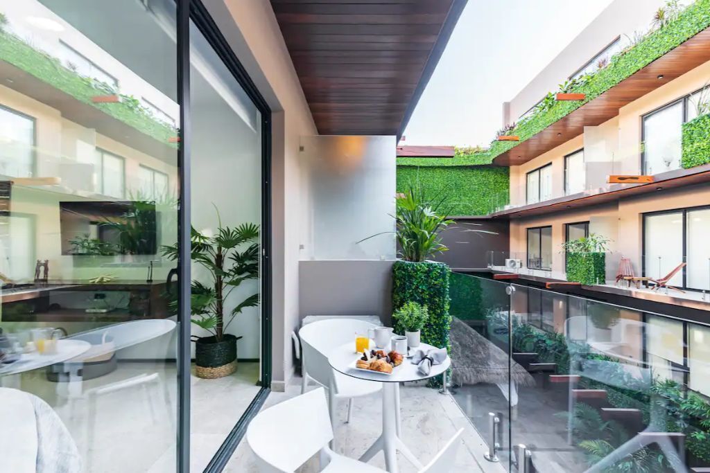 A modern outdoor dining area with a glass balustrade, potted plants, and a view of the building's interior.