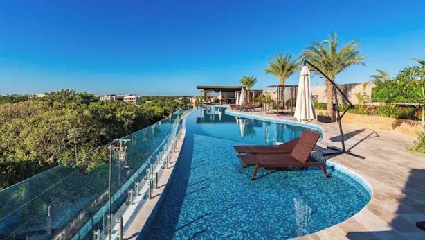A swimming pool with a glass barrier, lounge chairs, palm trees, and a clear blue sky. - Wonderful studio in Menesse Condominium (Tulum)