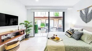 A modern hotel room with a bed, a flat-screen TV, a wooden console table, a plant, and a balcony with outdoor seating. - Wonderful studio in Menesse Condominium (Tulum)