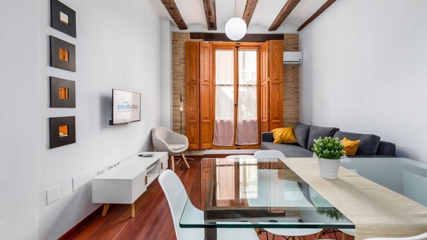 A modern living room with a glass dining table, wooden beams, a white sofa, and a potted plant.