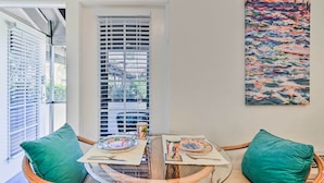 Een eetgedeelte met een glazen tafel, rieten stoelen en uitzicht door een raam met jaloezieën.