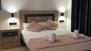 Een net opgemaakt bed met roze beddengoed en kussens, een nachtkastje met een lamp, en een wandlamp.