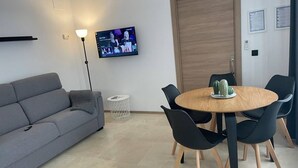 Een moderne woonkamer met een grijze bank, een ronde houten tafel met stoelen, een aan de muur bevestigde televisie en een cactus op de tafel.