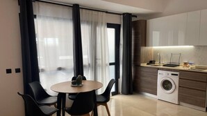 Een moderne keuken met een ingebouwde wasmachine, een eethoek met een ronde tafel en stoelen, en grote ramen voorzien van transparante gordijnen.