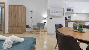 Une chambre d’hôtel moderne avec une armoire en bois, un lit recouvert d’une courtepointe bleue, une table à manger avec des chaises, un coin cuisine et un panneau encadré accroché au mur.