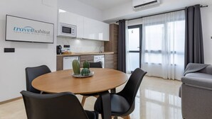 Une cuisine moderne avec des meubles blancs, un four à micro-ondes et une machine à café. On y trouve un coin repas avec une table ronde et des chaises noires. Un canapé gris est visible au fond.