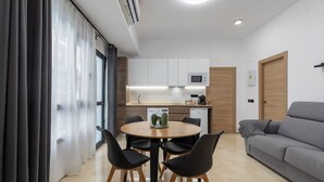 Una sala de estar moderna con un sofá gris, una mesa de comedor redonda de madera con cuatro sillas, y una zona de cocina con armarios blancos y un microondas.
