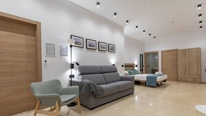 A modern hotel room with a grey sofa, a light blue armchair, a bed, a wardrobe, and a mirror.