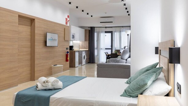 A modern hotel room with a neatly made bed, a small kitchenette, a seating area, and a view of the adjacent room.