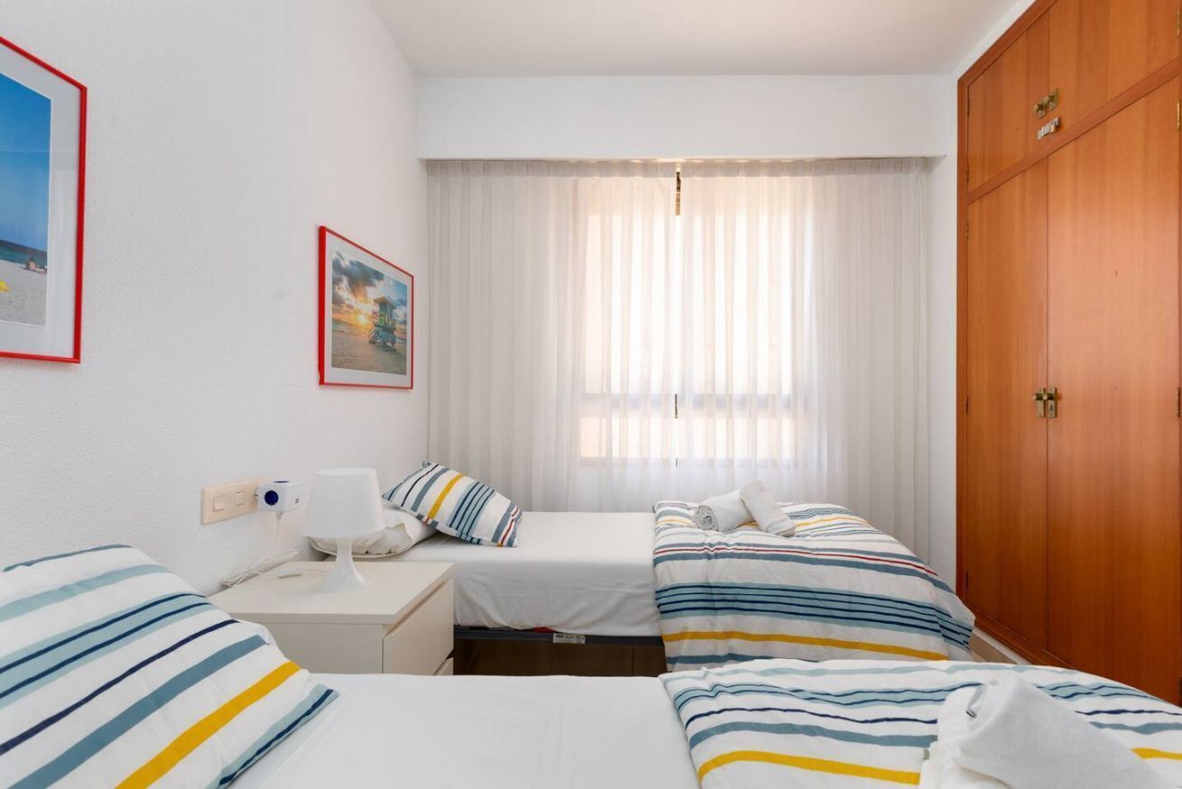 Habitación de hotel con dos camas, un armario de madera, una mesita de noche con lámpara y una ventana con cortinas traslúcidas.