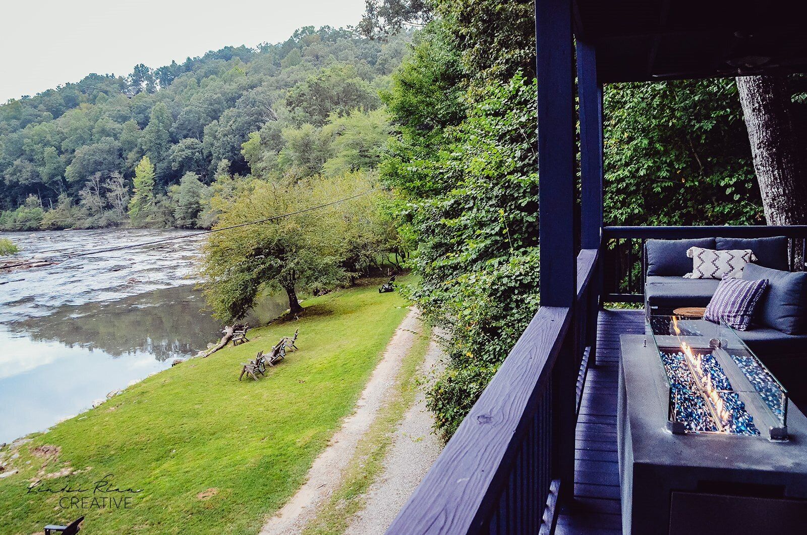 A riverside deck with a fire pit and outdoor seating, overlooking a river and forested hills.