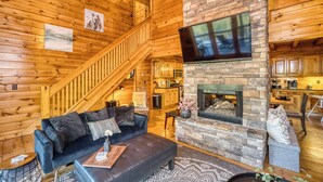 A cozy log cabin interior with a stone fireplace, wooden walls, a staircase, and a flat-screen TV mounted above the fireplace.
