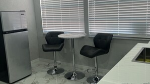 Eine moderne Küche mit Kühlschrank, zwei Barhocker, einem runden Tisch und großen Fenstern mit Jalousien.