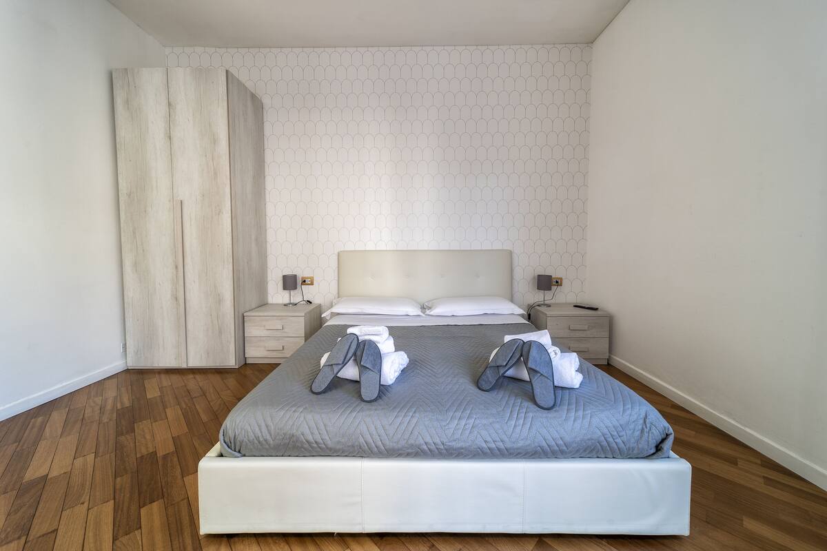 A neatly made bed with a grey quilt, two bedside tables with lamps, and a wooden wardrobe.