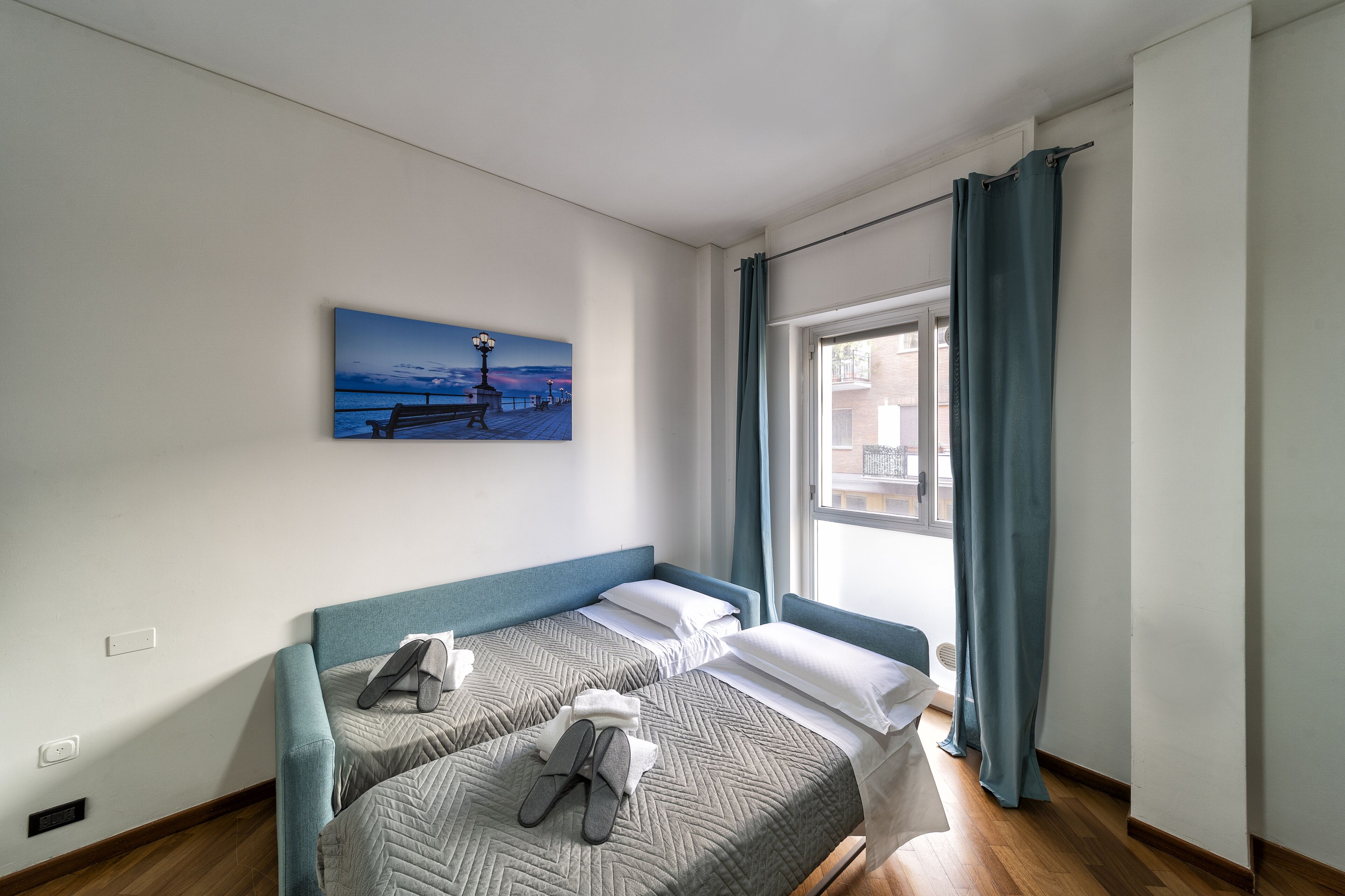 A room with two beds, a painting of a pier, and a window with curtains.