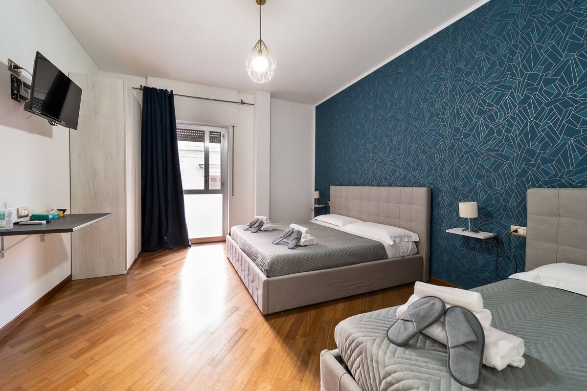 A modern hotel room with two beds, a flat-screen TV mounted on the wall, a bedside table with a lamp, and a large window with curtains.