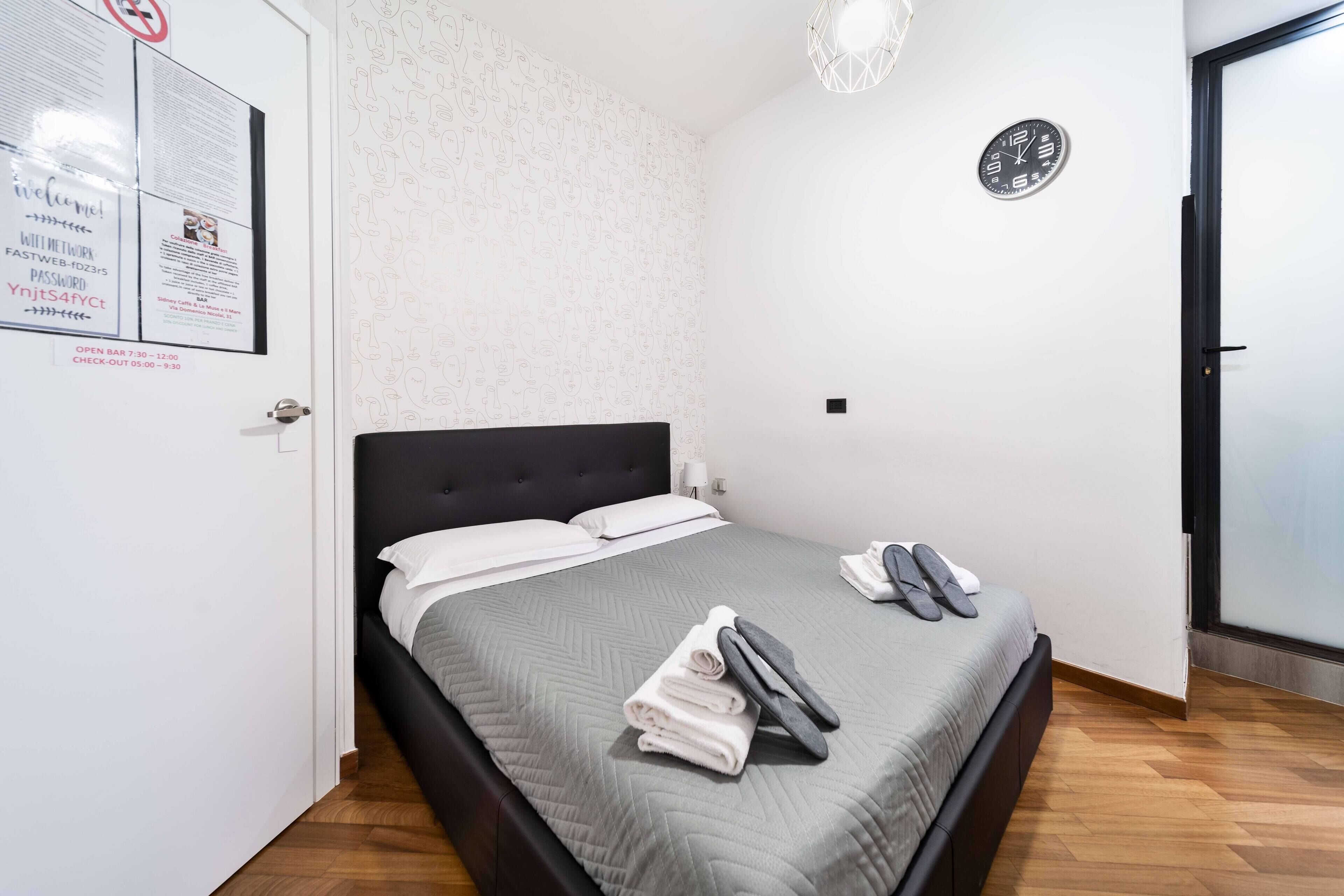 A neatly made bed with a grey quilt, white pillows, and a folded towel and slippers on the side.