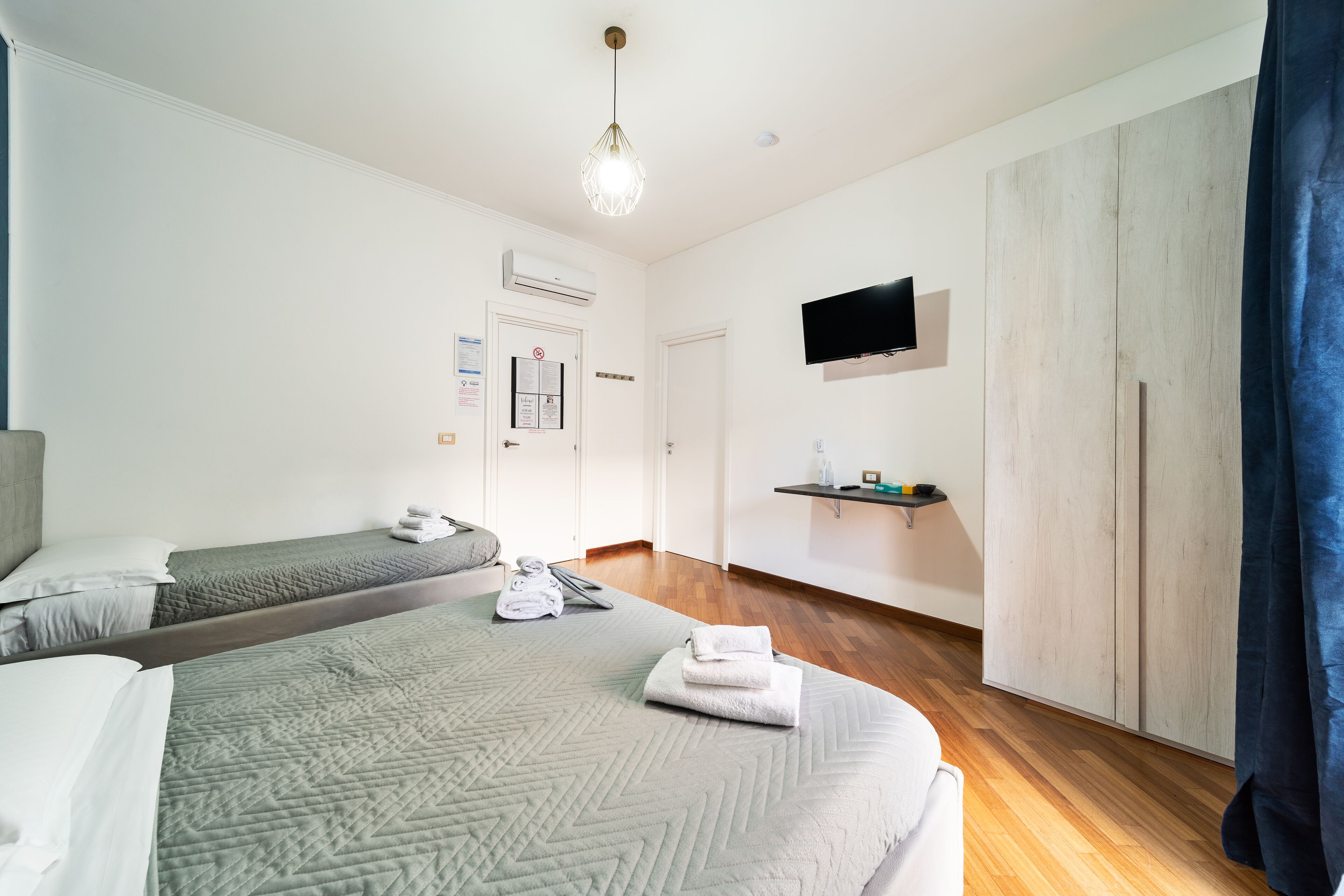 A hotel room with two beds, a TV, a wooden wardrobe, and a mounted shelf.