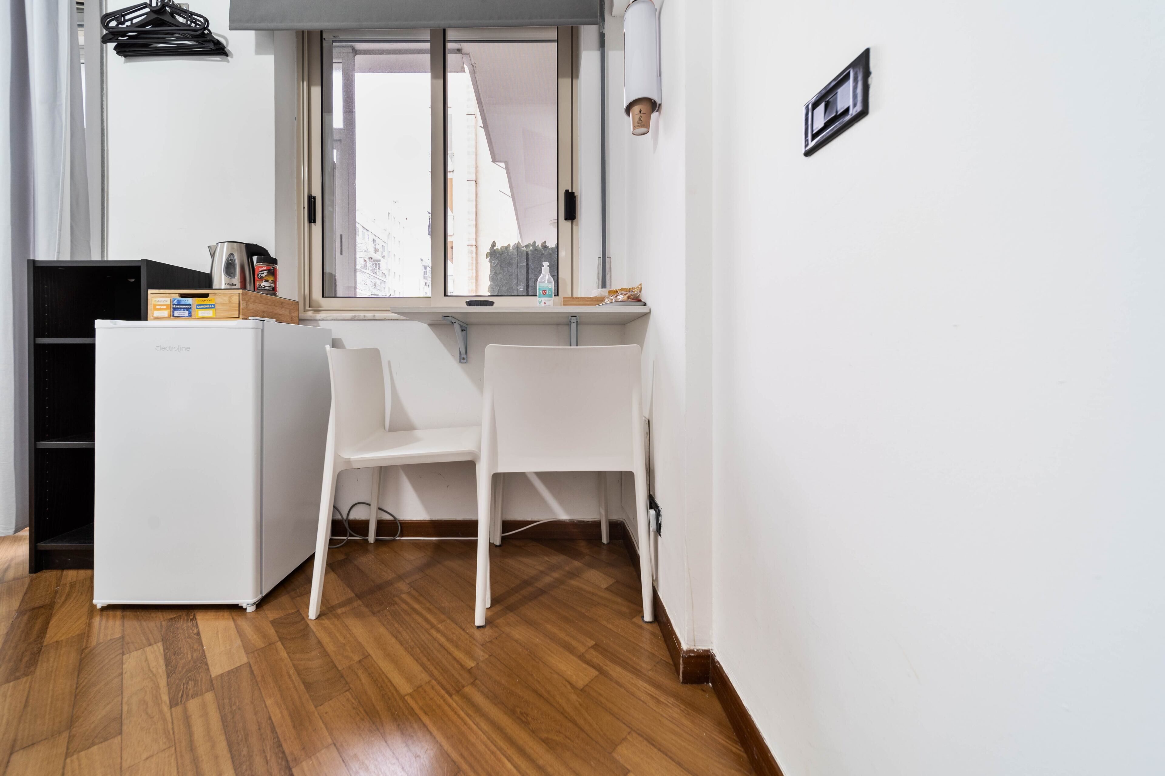 A compact kitchen area with a refrigerator, two white chairs, a window with a view of buildings, and a small countertop with items.