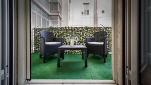 Une vue à travers la fenêtre d’une pièce où se trouvent deux fauteuils en osier, une petite table et un sol recouvert d’un tapis d’herbe artificielle verte.