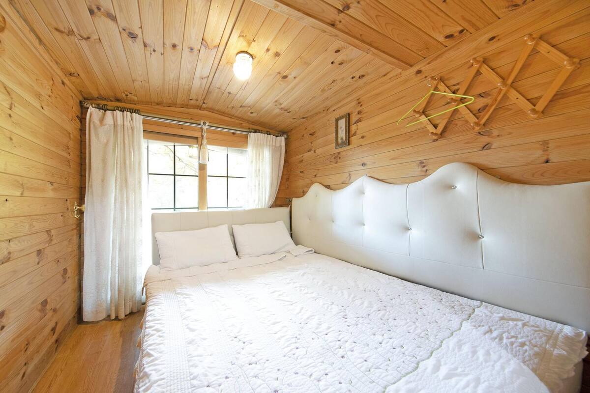 A wooden cabin bedroom with a large bed, wooden walls, and a window with curtains.