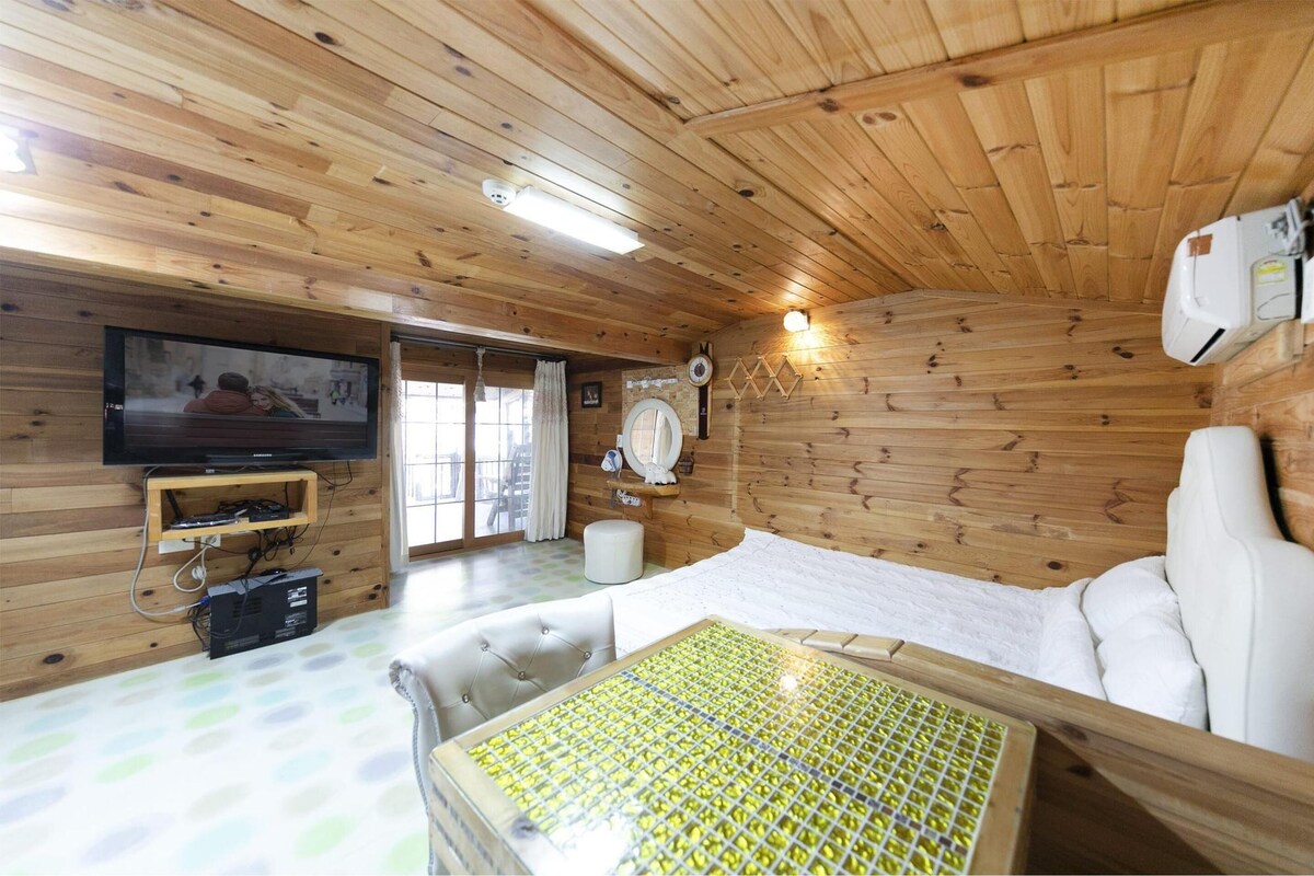 A wooden cabin room with a bed, a television, a heater, and a window with curtains.