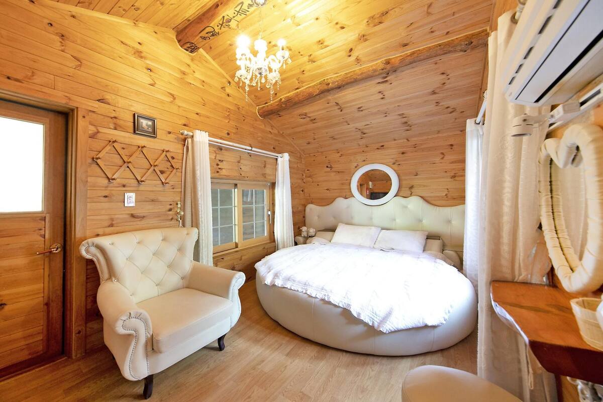 A wooden cabin bedroom with a round bed, a tufted armchair, and a chandelier.