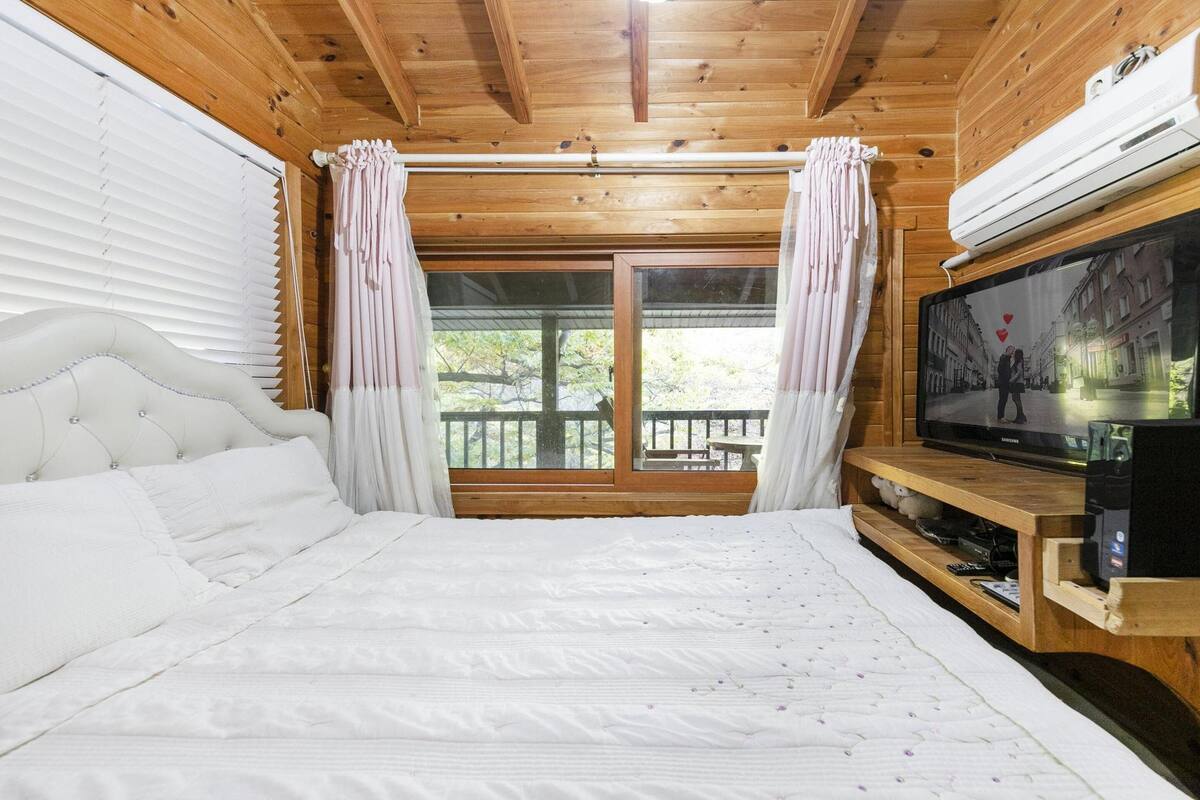 A bedroom with a bed, curtains, a television on a wooden stand, and a window with a view of the outdoors.