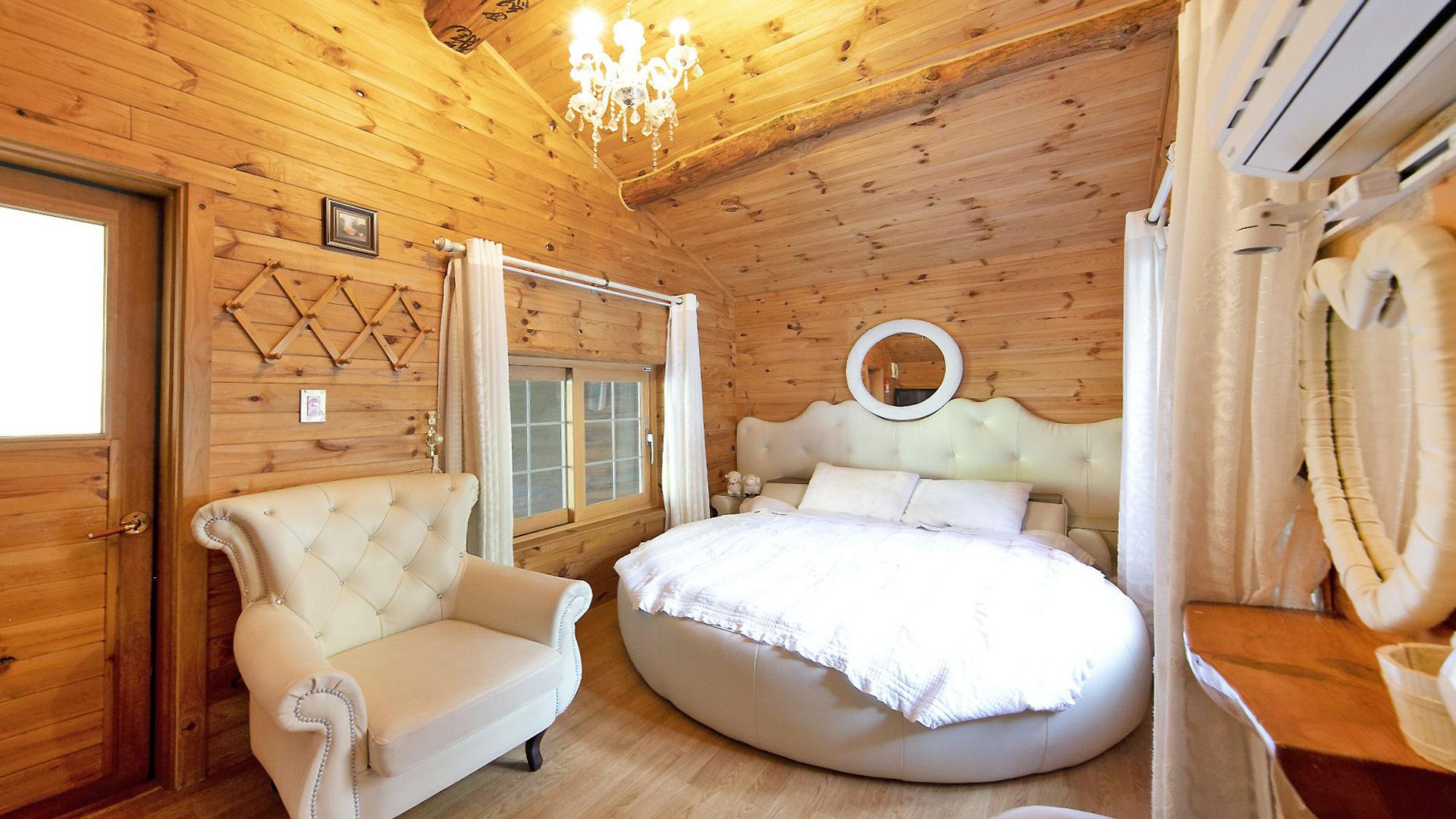 A wooden cabin bedroom with a round bed, a tufted armchair, and a chandelier.