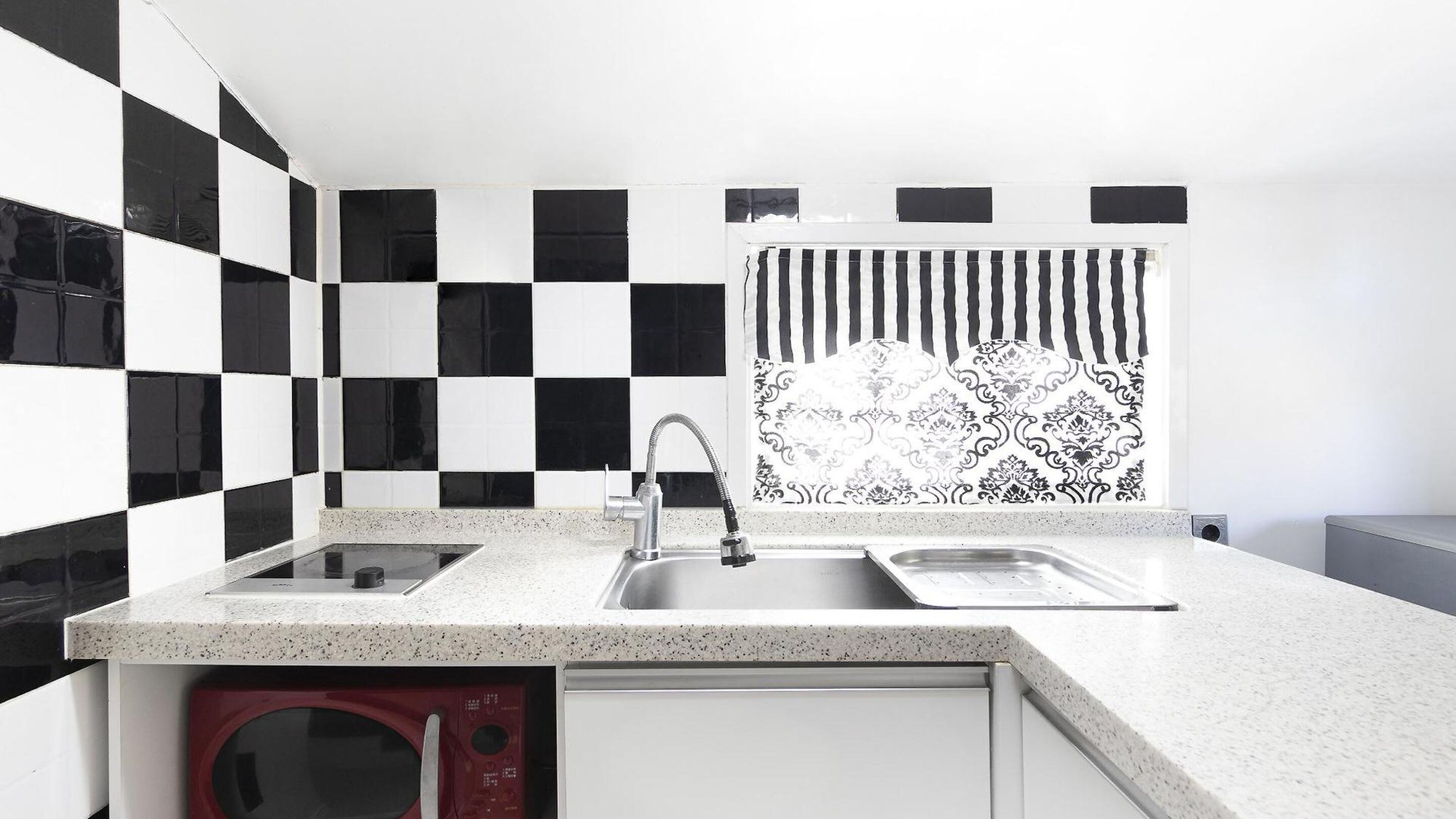 A kitchen with a checkered backsplash, a double sink, a microwave, and a patterned window treatment.