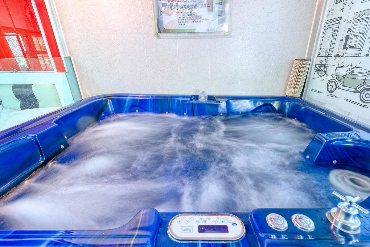 A hot tub with a control panel, surrounded by a tiled wall and a window with red curtains.