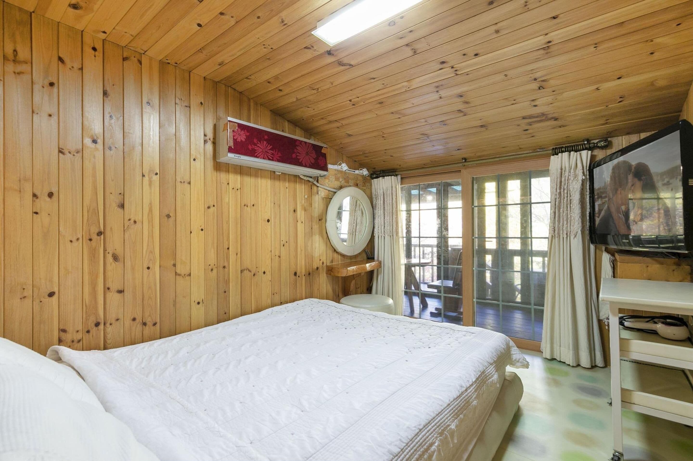 A wooden-paneled room with a bed, a TV, a mirror, and a window with curtains.