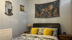 A bedroom with a patterned bedspread, a large tapestry, a mirror, and a bedside table with a lamp. - Beautiful Town Centre Apartment With River Terrace (Macclesfield)